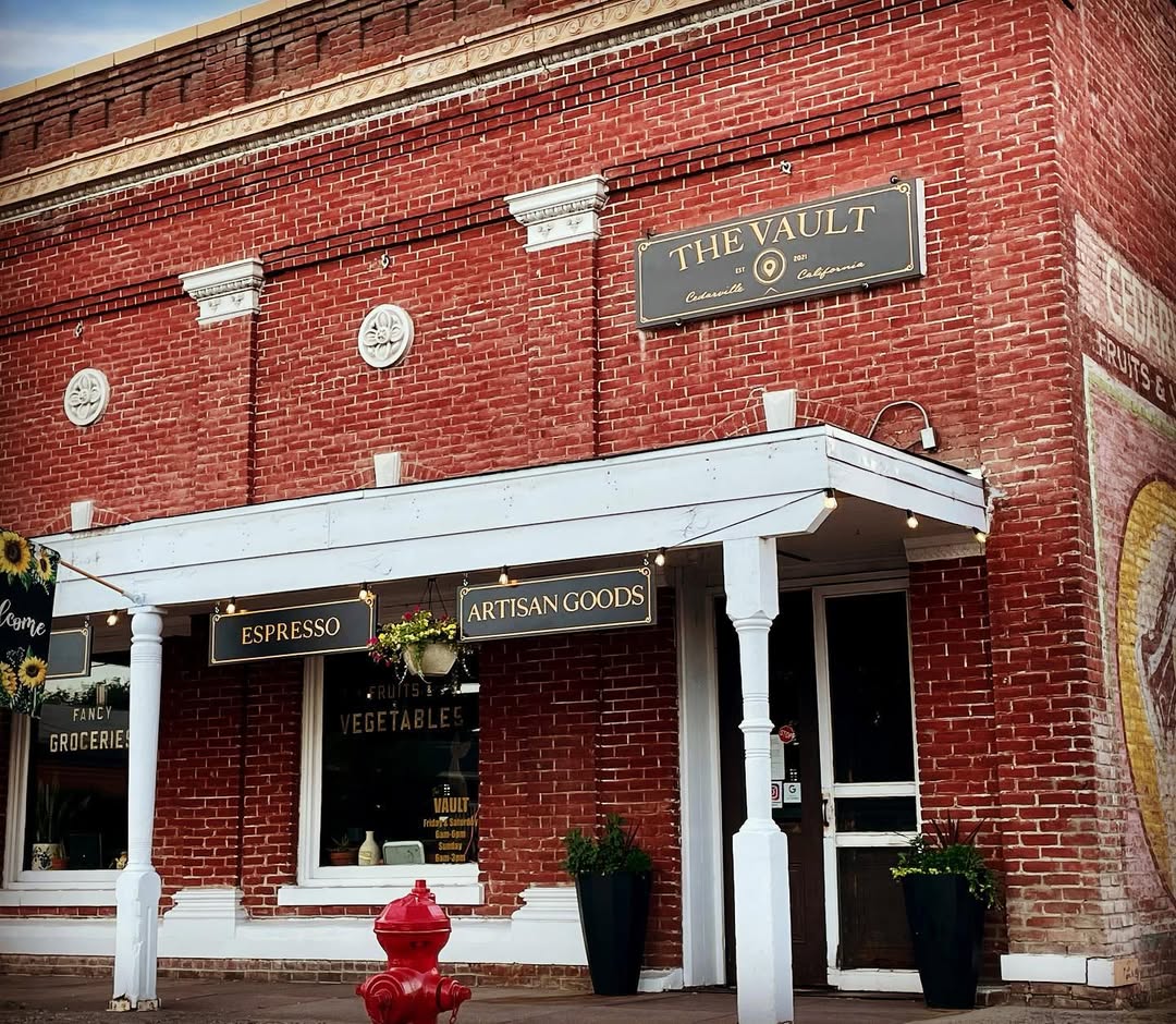 Brick storefront of The Vault coffee house in Cedarville, California, with hanging signs reading 'ESPRESSO' and 'ARTISAN GOODS' and a vintage red fire hydrant out front